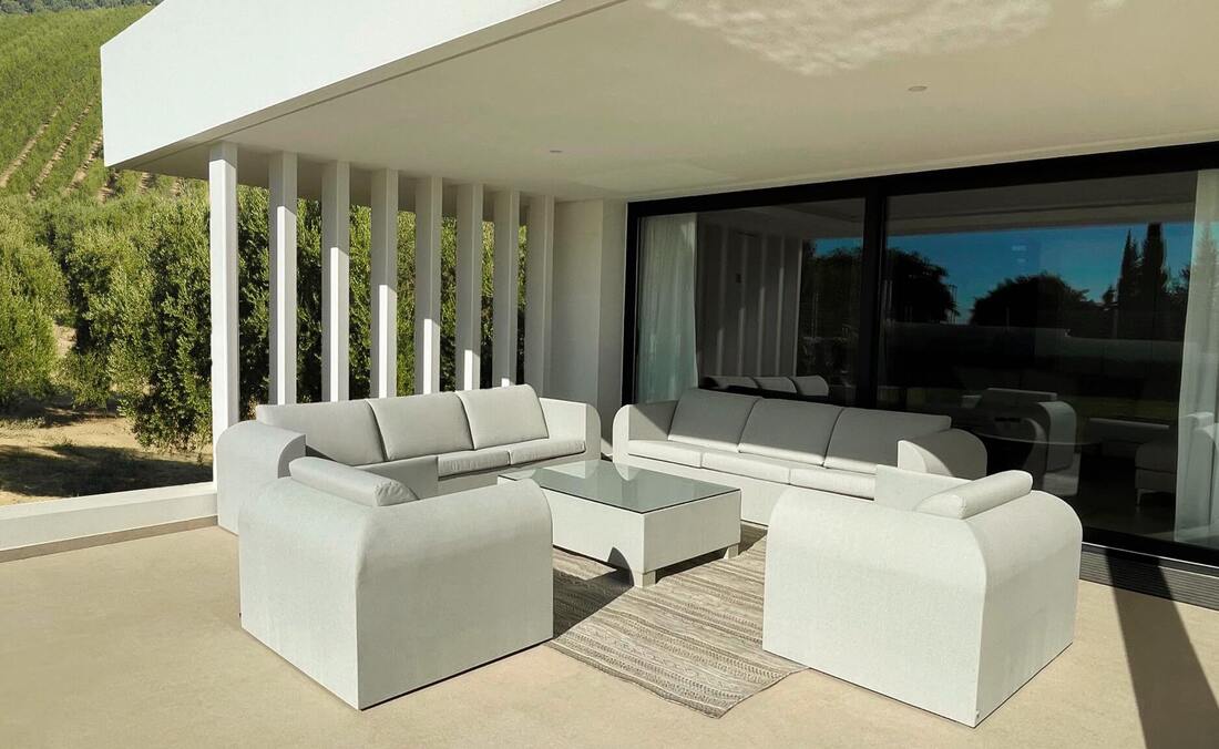 Porche en una casa moderna de Marbella con dos sofás de color lino, dos sillones, una mesa de centro y una alfombra para exterior de color arena con fondo de campo de olivos